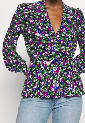 Frau trägt eine langärmelige Wickelbluse mit Blumenmuster in Lila, Blau, Weiß und Grün, kombiniert mit hellblauen Jeans.