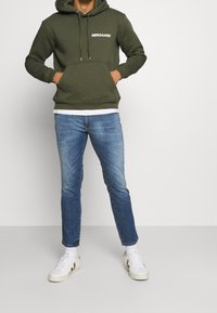 Olive green hoodie with front pocket and drawstrings, white logo. Worn with light blue jeans and white sneakers. Minimalist design.