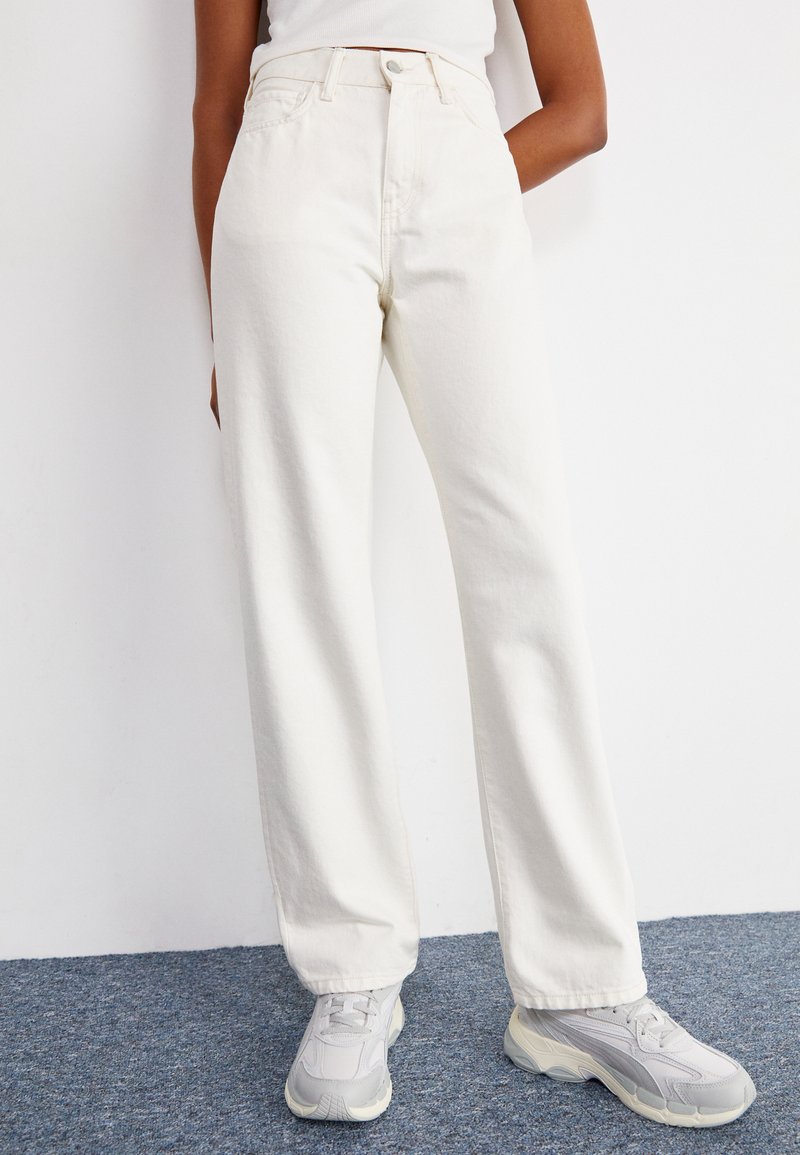 White cotton wide-leg jeans with stitched pockets, high waist, and a smooth finish. Paired with light gray sneakers.