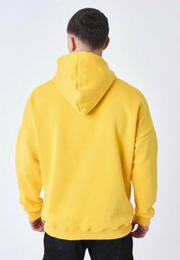 Sudadera amarilla con gran cuello de embudo, corte oversize, textura suave y puños acanalados. Cuenta con un bolsillo canguro en la parte frontal.