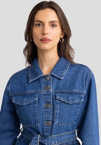 Jeans jakna z pasom, dvema žepoma na prsih in gumbi. Ima klasičen ovratnik in modro pranje z vidnimi šivi.