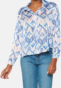 Camicia con bottoni e maniche lunghe, caratterizzata da un motivo geometrico blu e rosa su un tessuto traslucido e testurizzato. Design con orlo ondulato.