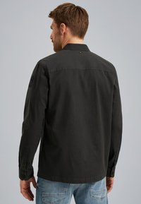Chemise à manches longues vert foncé avec un col, des poignets boutonnés et un détail subtil de logo sur la manche. Texture en coton.