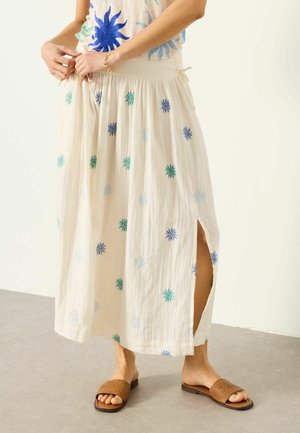 Frau richtet cremefarbenen Maxirock mit bunter Blumenstickerei und seitlichem Schlitz, trägt geflochtene braune Pantoletten.