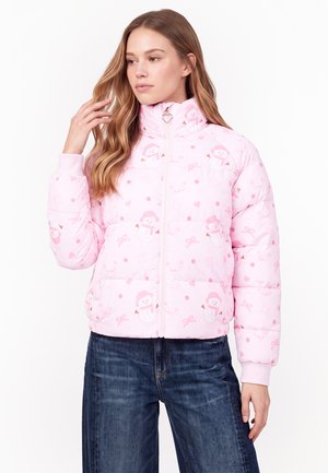 Ung kvinde iført en lyserød dunjakke med snemandsmønstre og mørkeblå jeans, stående mod en ensfarvet hvid baggrund.