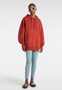 Roter Pullover-Hoodie mit Kordelzug und Fronttasche. Kombiniert mit hellblauen Leggings und gemusterten Schlupfschuhen. Weiße Sonnenbrillen runden den Look ab.