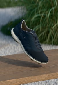Schwarzer Slipper mit einer glatten synthetischen Oberseite, strukturierten elastischen Schnürsenkeln und einer weißen Sohle, die über einer Holzfläche schwebt.