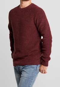 Maglione lavorato a maglia bordeaux con texture a costine, scollatura rotonda e maniche raglan, abbinato a jeans azzurri chiari.