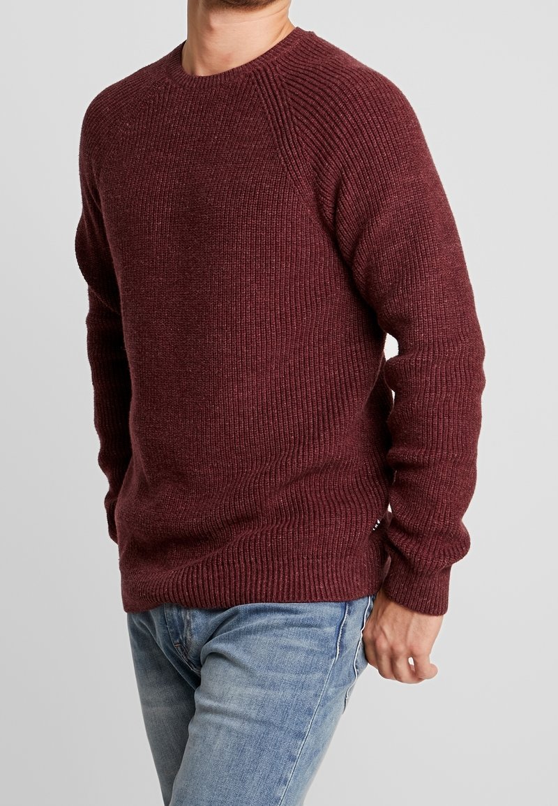Maglione lavorato a maglia bordeaux con texture a costine, scollatura rotonda e maniche raglan, abbinato a jeans azzurri chiari.