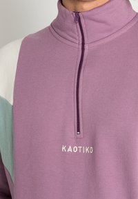 Fialový fleecový pulovr s polozipem, s akcenty v světle modré a bílé barvě; obsahuje vyšitý logo "KAOTIKO". Měkká textura, ležérní design.