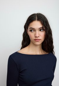 Temno modra rebrasta majica z dolgimi rokavi in širokim izrezom v obliki čolna, zasnovana je oprijeto in ima gladko teksturo. Preprost in čist videz.