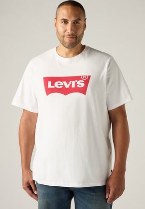 Mężczyzna w białym t-shircie Levi's z czerwonym logo, stojący na jednolitym jasnym tle.