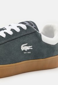 Baskets en daim gris foncé avec lacets blancs, semelle en caoutchouc naturel, et un logo de crocodile sur le côté. Dotées d'un renfort au talon contrastant et d'une surface texturée.