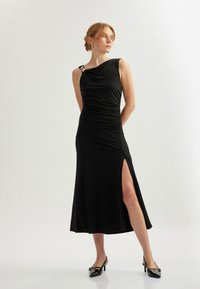Schwarzes, ärmelloses Kleid mit Schalkragen, gerafften Seiten und einem hohen Schlitz bis zum Oberschenkel. Verfügt über goldfarbene Akzentbeschläge an der Schulter. Glatte Textur.
