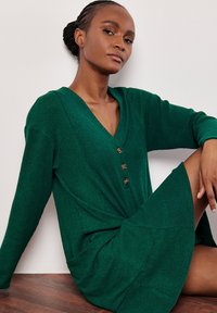 Abito verde a coste con scollatura a V, maniche lunghe e bottoni in legno. Presenta un nastro in vita e tasche laterali. Tessuto liscio e vestibilità comoda.
