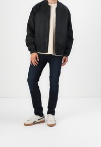 Svart bomberjacka med dragkedja, lättviktsmaterial, ribbade ärmslut, bärs över en beige t-shirt, kombinerad med mörka jeans och krämfärgade sneakers.