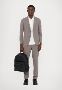 Grauer maßgeschneiderter Anzug mit geknöpftem Blazer, weißem Poloshirt, schwarzem Rucksack in runder Form und geschnürten schwarzen Schuhen. Glatte Stoffstrukturen.