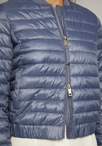 Personne portant une veste matelassée bleu brillant avec deux fermetures éclair argentées apparentes, l'une partiellement fermée, l'autre complètement ouverte.