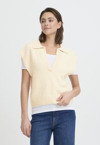 Gilet en maille jaune clair avec un col en V, des manches courtes et un ourlet côtelé, superposé sur une chemise blanche et associé à un jean en denim.