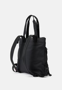 Sac à dos noir en forme de carré avec des bretelles rembourrées et deux longues poignées de transport, présenté sur un fond blanc uni.