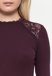 Paarse top met lange mouwen, gemaakt van ribstof, met kantaccenten op de schouderranden en een golvende halslijn.