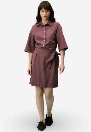 Femme portant une robe mauve ceinturée mi-longue avec boutons, manches mi-longues, et mocassins noirs, debout devant un fond uni.
