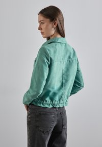 Veste en velours côtelé teal avec un col, un ourlet élastique et deux poches poitrine, associée à un pantalon en denim gris foncé.