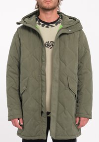 Olijfgroen gewatteerd jack met een capuchon, voorzien van drukknoopsluitingen, zijzakken en een zachte textuur. Daaronder is een beige shirt met een patroon zichtbaar.