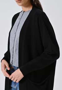 Eine Frau trägt eine schwarze, offene Strickjacke über einem geknöpften grauen Oberteil und blauen Jeans. Sie hält die Kanten der Strickjacke auf Taillenhöhe.