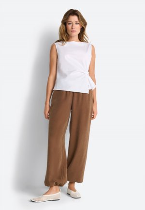 Naine seistes valge seina taustal, seljas varrukateta valge top, millel on küljesõlm, laiad pruunid püksid ja valged kootud kingad.