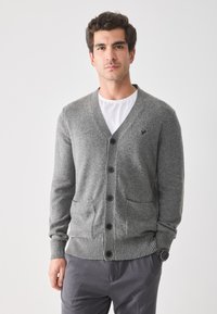 Cárdigan gris con escote en V, cinco botones negros, puños y dobladillo acanalados, con dos bolsillos frontales, llevado sobre una camiseta blanca.