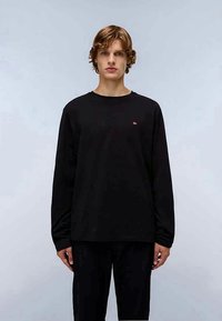 Jovem com cabelo castanho ondulado está de frente, vestindo uma camiseta preta de mangas longas e calças pretas, contra um fundo cinza claro.