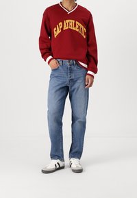 Sudadera burdeos con "GAP ATHLETIC" en amarillo, puños y cuello de canalé blancos. Jeans rectos de mezclilla azul claro y zapatillas blancas con rayas negras.