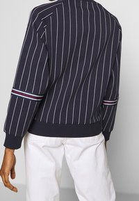 Sudadera azul marino con rayas verticales blancas, puños y dobladillo acanalados, y rayas de acento multicolores en las mangas. Vista desde atrás.