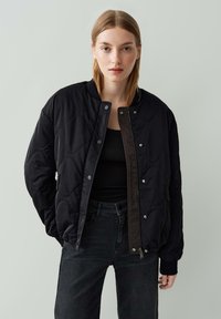 Jeune femme aux cheveux blonds raides portant une veste matelassée noire, un haut noir et un jean foncé, le tout sur fond gris clair uni.