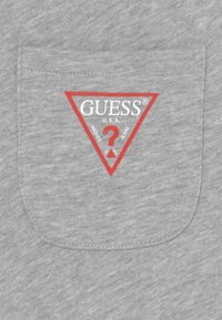 Grå stoflomme med det røde og hvide "GUESS" logo inden for et trekantet design. Baggrundens tekstur er meleret.