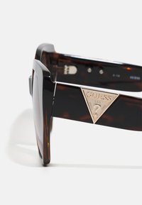 Ochelari de soare cu model broască țestoasă, cu o ramă geometrică neagră, având un accent metalic în formă de triunghi pe lateral. Lentilele sunt întunecate și reflectorizante.