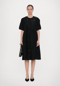 Black knee-length dress with short sleeves, textured fabric featuring floral cutouts. Paired with a dark green croc-patterned handbag and black heels.