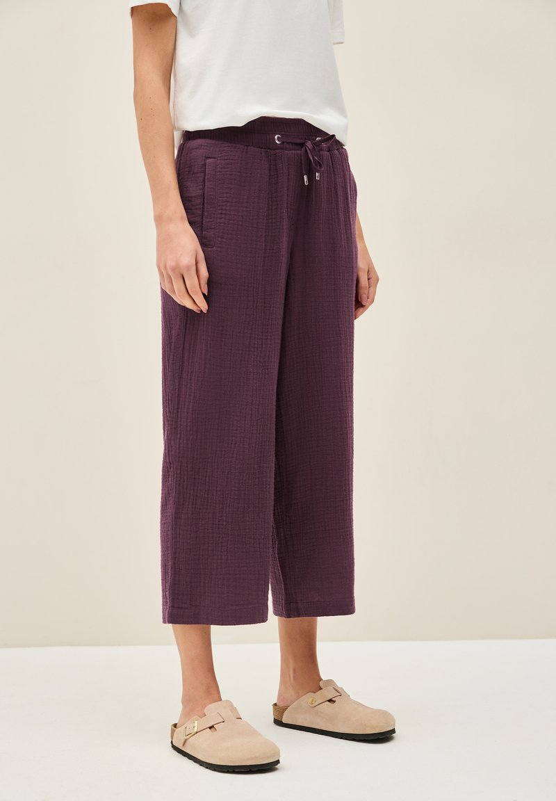 Pantalons courts texturés violets avec une taille à cordon de serrage et des poches latérales, associés à des sabots en suede légers et à enfiler.