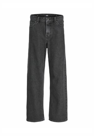 Ragazzo in piedi e sorridente, indossa una giacca beige sopra una camicia blu a quadri, pantaloni grigio scuro e scarpe marrone chiaro, su uno sfondo uniforme.