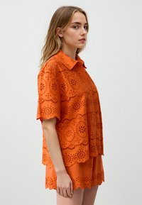 Oranje oogjes top met een gegolfde zoom en korte mouwen, met decoratieve uitsparingen en een kraaghalslijn. In combinatie met bijpassende shorts.