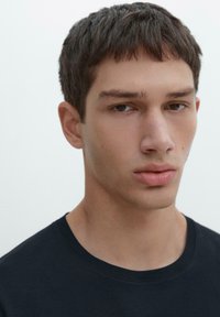 Giovane uomo con capelli corti e scuri, espressione neutra, che indossa una maglietta nera a girocollo, su uno sfondo chiaro e semplice.