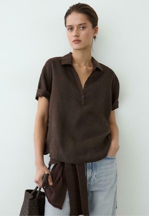 Jeune femme aux cheveux courts bruns portant une chemise marron foncé à col, un jean bleu clair et tenant un sac à main tressé marron foncé.