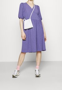 Robe violette à pois avec manches courtes bouffantes, associée à un petit sac blanc porté en bandoulière et des baskets grises avec des accents jaunes.