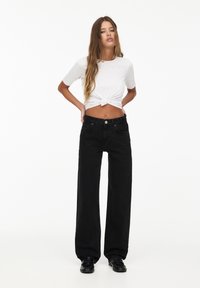 Zwarte wijde jeans met voorkruizen, gecombineerd met een kort, geknoopt grijs t-shirt. De stof is soepel en iets getailleerd bij de taille.