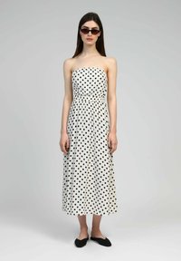 Robe blanche sans bretelles avec des pois noirs, taille cintrée, silhouette fluide et ourlet longueur cheville. Associée à des ballerines noires.