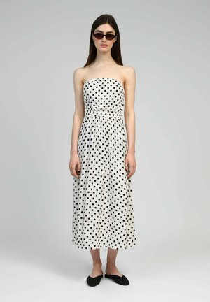 Robe blanche sans bretelles avec des pois noirs, taille cintrée, silhouette fluide et ourlet longueur cheville. Associée à des ballerines noires.