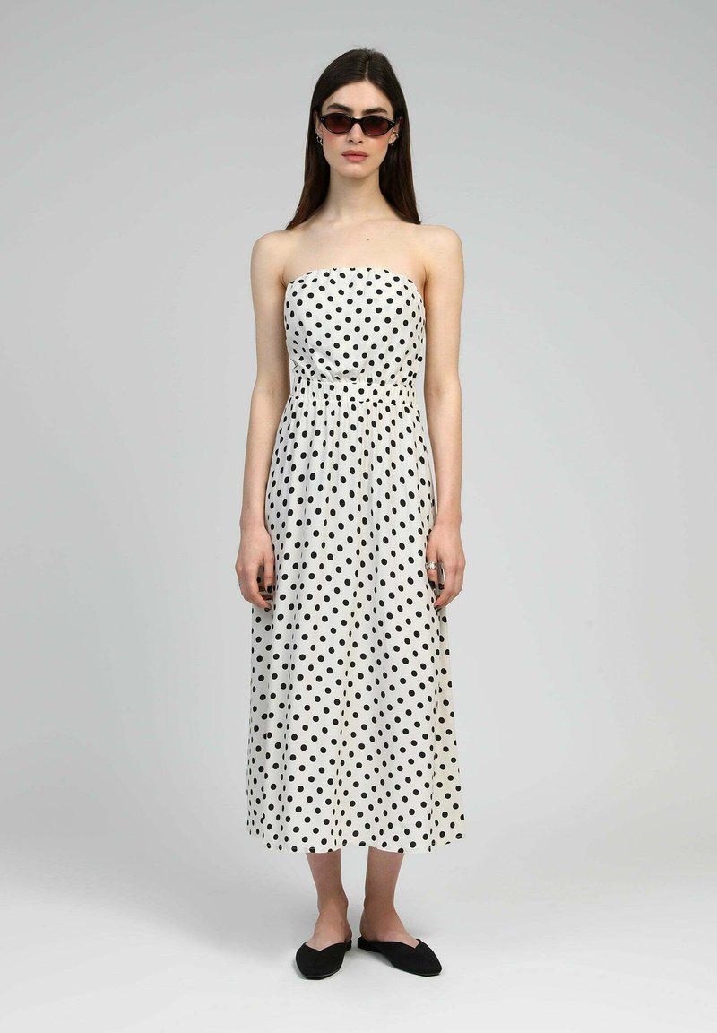 Robe blanche sans bretelles avec des pois noirs, taille cintrée, silhouette fluide et ourlet longueur cheville. Associée à des ballerines noires.