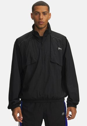 Junger Mann trägt eine schwarze Under Armour Pullover-Jacke mit halbem Reißverschluss und elastischen Bündchen, steht vor einem schlichten weißen Hintergrund.