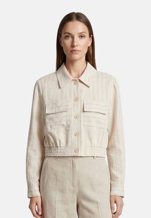 Donna con capelli castani lisci che indossa una camicia beige corta con bottoni e due tasche frontali abbinate a pantaloni a vita alta.